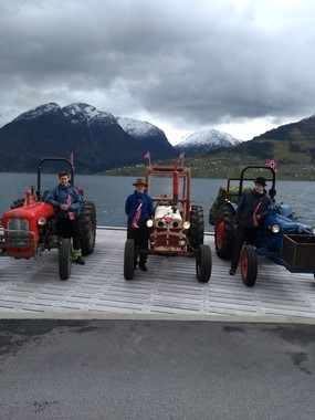 Tre eldre traktorar pynta med bjørkeris og norske flagg, og menn med fine hattar og flagg. Fjord og fjelllandskap i bakgrunnen.